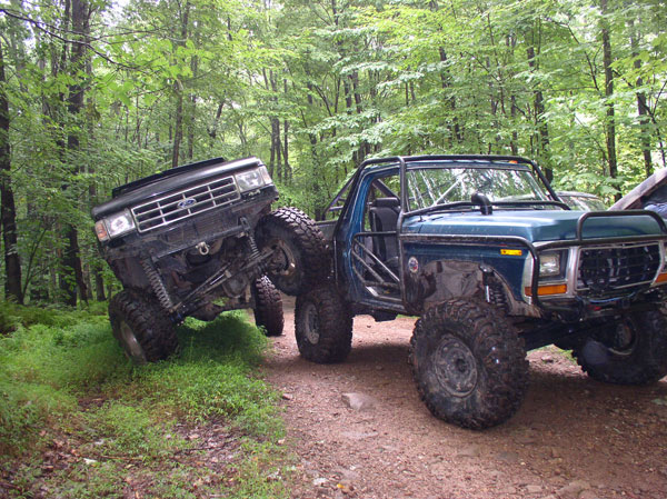 Modified blue 1979 Ford Bronco and black Ford truck off-roading in woods