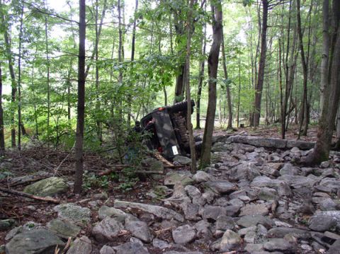 1988 Ford Bronco - An overturned vehicle lies on its side amidst rocks and trees in a forest clearing.