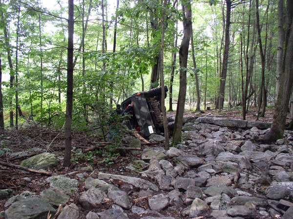 Overturned dark-colored off-road vehicle on its side in a rocky, wooded area