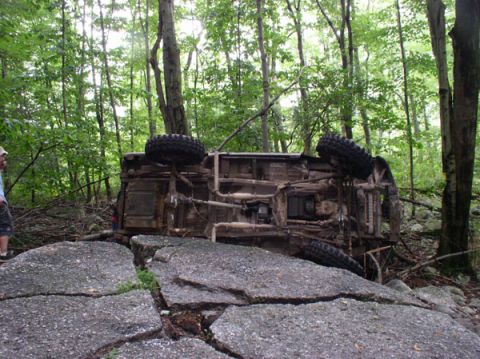 1988 Ford Bronco - A vehicle lies overturned on large rocks in a wooded area, with a person observing in the background.