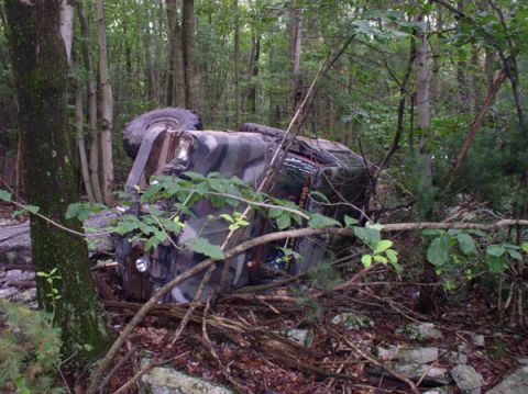 1988 Ford Bronco - An overturned camouflage SUV rests on its side amidst dense forest foliage.