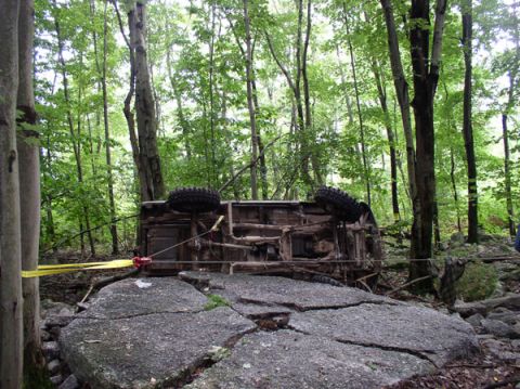 1988 Ford Bronco - A 4x4 lies rolled onto its side on a rocky wooded trail, showing the frame rails, mud-caked underbody, exhaust and mud-terrain tires. A yel…