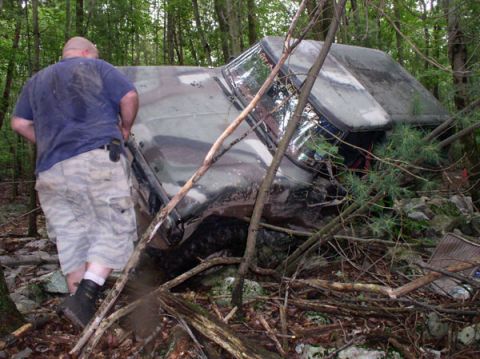 1988 Ford Bronco - A person inspects a camouflaged vehicle that has gone off-road and is entangled in trees and branches.