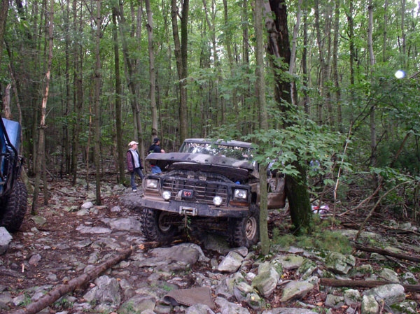 Lifted hoodless Ford Bronco off-camber on a rocky wooded trail with spectators