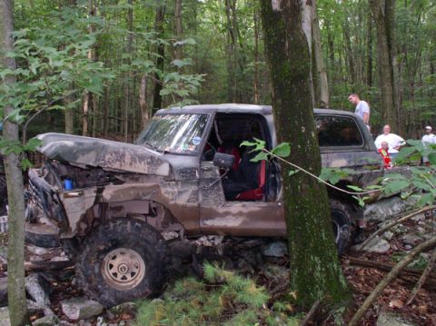 1988 Ford Bronco - A mud-caked, doorless full-size Bronco-style 4x4 is winched over a rocky ledge in the woods, its cable running past a large tree and the fr…
