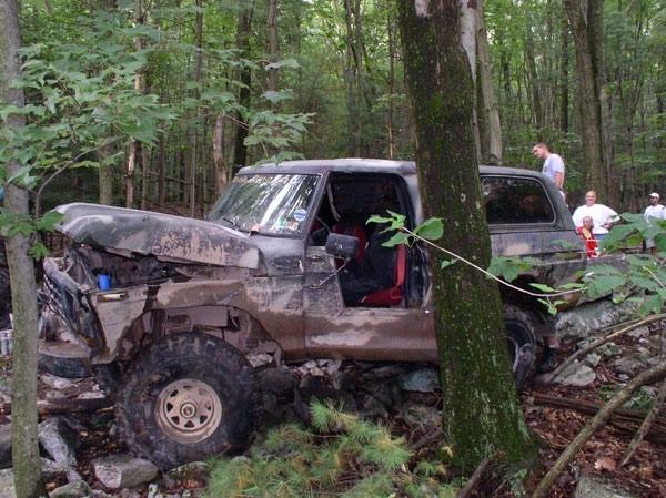 Mud-covered doorless 4x4 with crumpled front end winching over rocks in the woods