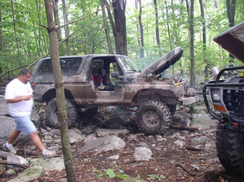 1988 Ford Bronco - Two off-road vehicles are seen in a wooded area with one Bronco having its hood up.
