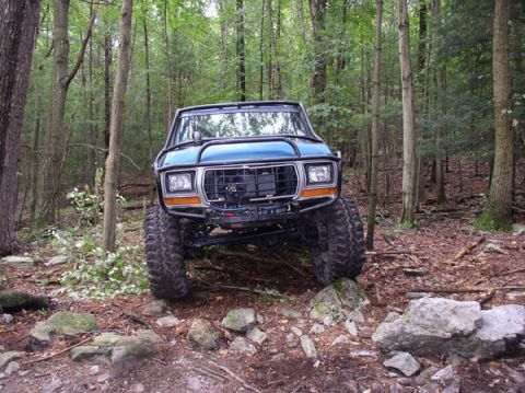 1988 Ford Bronco - Modified SUV with off-road tires driving over rocks in a wooded area.