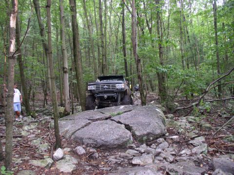 1988 Ford Bronco - A black Ford Bronco navigates a rocky trail through a dense forest, with another vehicle visible in the background.