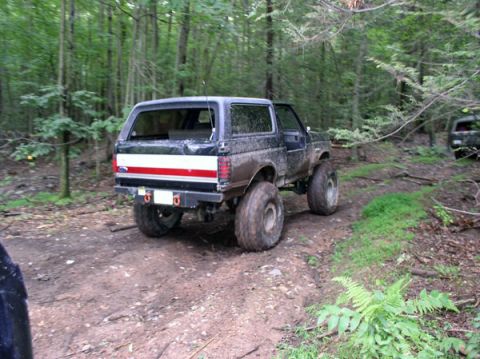 1988 Ford Bronco - A lifted Ford Bronco navigates a wooded trail, kicking up dirt. Another vehicle is parked further down the path.