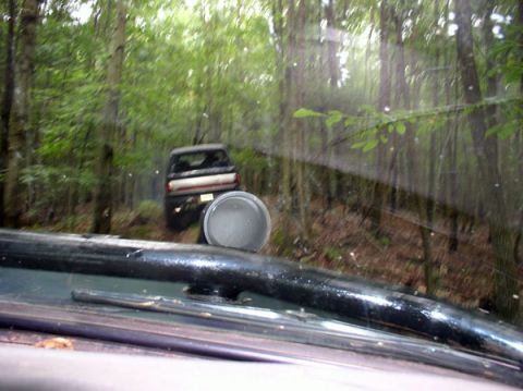 1988 Ford Bronco - A dark SUV navigates a wooded trail, viewed through the windshield of another vehicle.