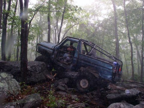 1988 Ford Bronco - A heavily modified off-road vehicle tackles a challenging rocky trail in a wooded area.