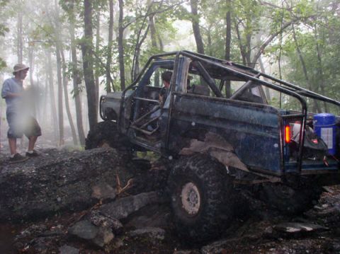 1988 Ford Bronco - Side view of a blue doorless, topless full-size 4x4 with a full exterior roll cage, cut fenders and a red fuel can plus blue jug strapped i…