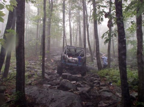 1988 Ford Bronco - An off-road vehicle navigates a challenging rocky path through a misty forest, with a person observing nearby.