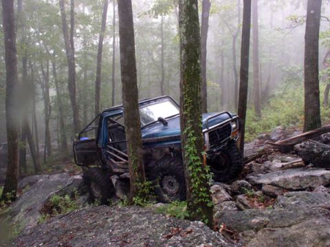 1988 Ford Bronco - Off-road truck with extensive modifications is stuck on a rocky trail in a forest.