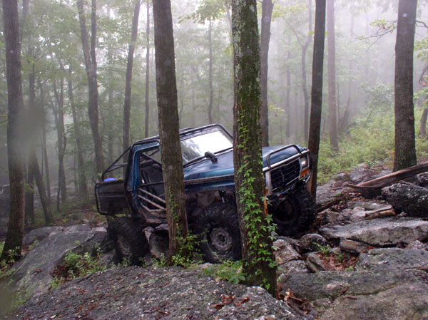 Modified blue truck with roll cage stuck between trees on rocky terrain