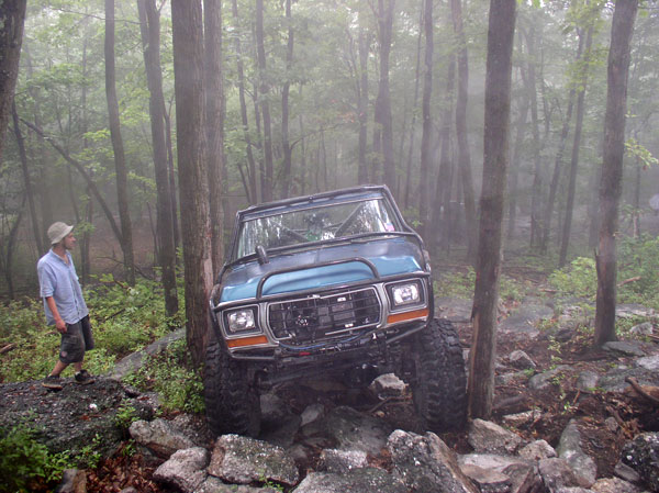 Blue off-road vehicle with large tires climbing over rocks in a forest. Person in hat watches
