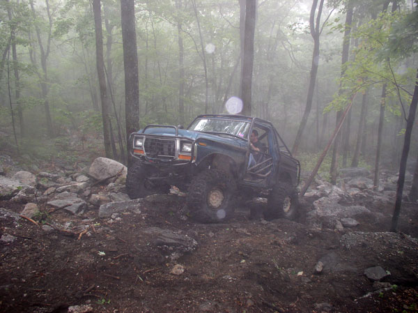 Ford Bronco modified for off-roading, driving through a rocky, wooded area