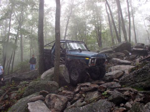 1988 Ford Bronco - Off-road vehicle with roll cage tackles a challenging rocky path in a wooded area.