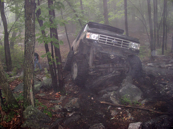 Ford Bronco climbing over rocks on a wooded trail. Grille looks like 1987