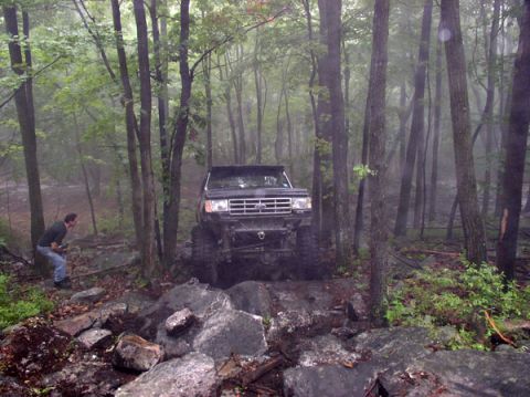 1988 Ford Bronco - A black 1987 Ford Bronco tackles a challenging off-road course amidst trees and rocks.