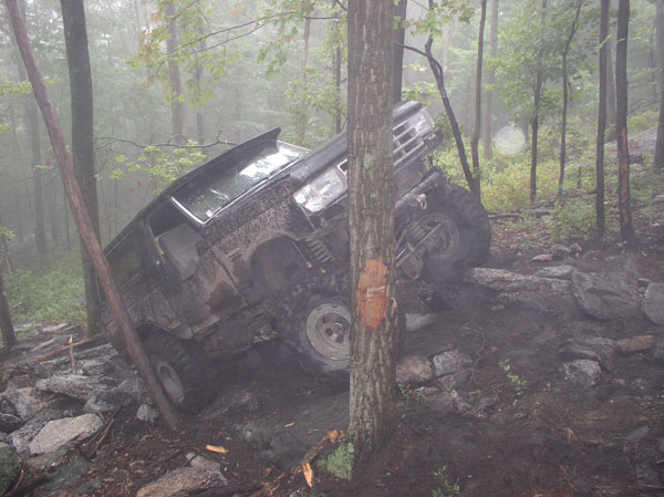 1988 Ford Bronco tilted on its side, stuck between rocks and trees on a muddy trail