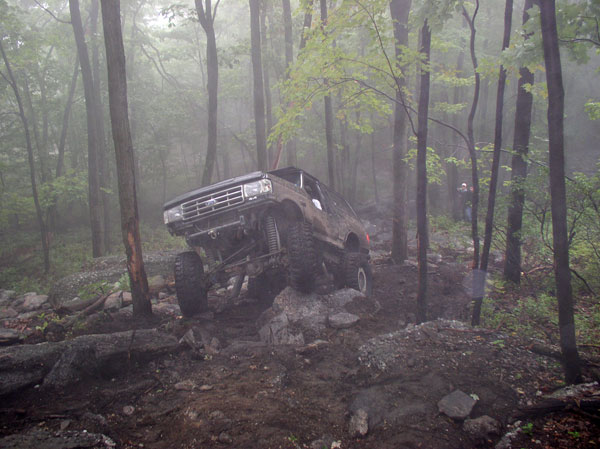 Brown 1987-1991 Ford Bronco off-roading on a rocky, wooded trail with lifted suspension