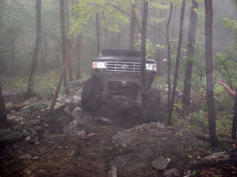 1988 Ford Bronco - A lifted Ford Bronco navigates a challenging, muddy, and rocky trail through a misty forest.