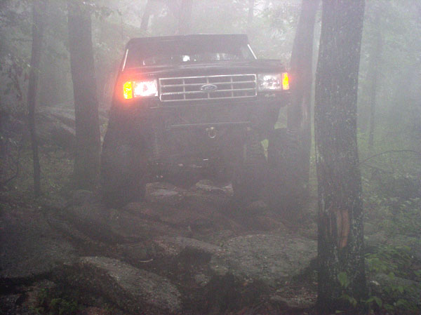 Ford Bronco driving over rocks in foggy woods. Grille indicates 1987-1991 model