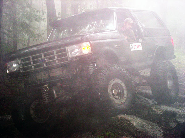 Ford Bronco climbing over rocks in foggy woods. Driver has a 'STAFF' sticker on the door