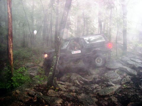 1988 Ford Bronco - Rear three-quarter view of a dark lifted Bronco-style SUV picking its way down a wet, rocky trail in heavy fog, brake light glowing and a w…