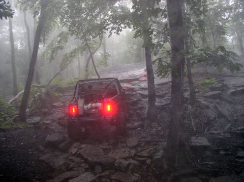 1988 Ford Bronco - An off-road vehicle with a roll cage navigates a challenging, rocky path through a misty forest.