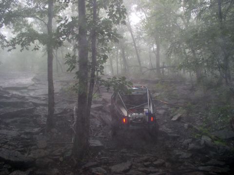 1988 Ford Bronco - A 1988 Ford Bronco navigates a challenging, rocky, and muddy off-road trail surrounded by dense trees.
