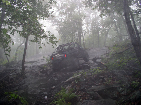 Off-road vehicle with roll cage climbing a rocky, wooded trail in fog