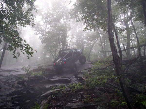 Dark off-road vehicle with a roll cage climbs a rocky, wooded incline on a foggy day