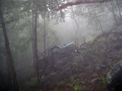 1988 Ford Bronco - A blue 1988 Ford Bronco is seen partially submerged in foliage and rocks on a steep, foggy incline, appearing stuck.