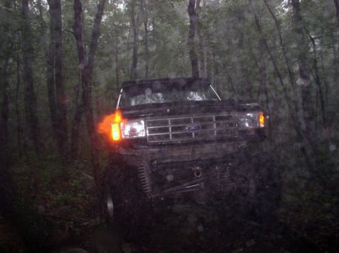 1988 Ford Bronco - A black Ford Bronco is seen off-roading through a dense forest with its headlights and turn signals illuminated.