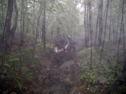 1988 Ford Bronco - A modified off-road vehicle descends a challenging, wet, and rocky trail through a dense forest.