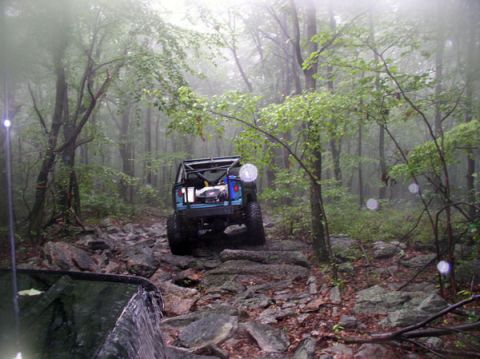 1988 Ford Bronco - A blue off-road vehicle navigates a challenging, rocky path through a dense, fog-shrouded forest.