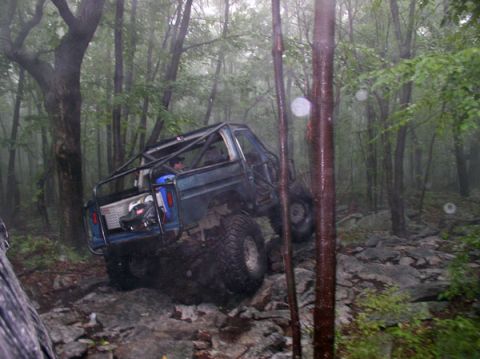 1988 Ford Bronco - Modified SUV with a prominent roll cage tackles a challenging rocky trail in a wooded area.