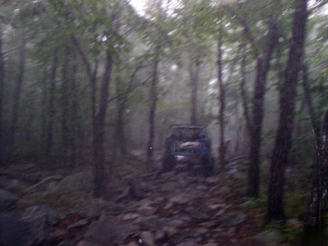 1988 Ford Bronco - A blue SUV navigates a rough, rocky path through a dense, misty forest.