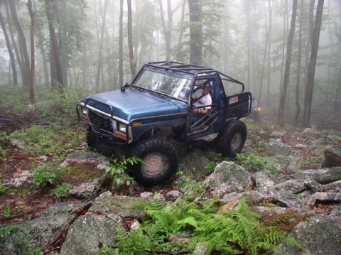 1988 Ford Bronco - A modified blue SUV drives through a misty forest on a rocky path.