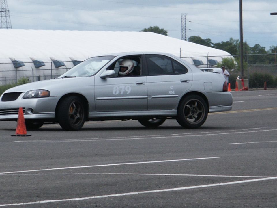 Silver sedan with racing number 879 on the door, driver wearing a helmet, on a track with orange cones