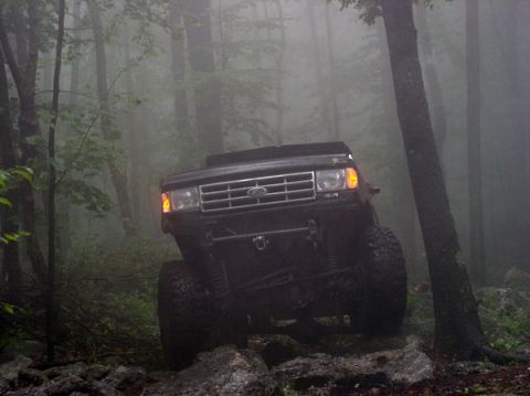 1988 Ford Bronco - Head-on view of a lifted dark Ford 4x4 cresting a wet rock ledge in foggy woods, amber marker lights glowing and the Ford grille and hood d…