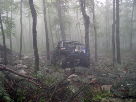 1988 Ford Bronco - Rear view of a blue chopped-top 4x4 buggy with an exposed roll cage, spare tire and bed-mounted gear crawling a jagged rock garden in foggy…