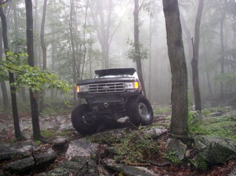1988 Ford Bronco - A lifted black Ford Bronco navigates a challenging, rocky path through a misty woodland.
