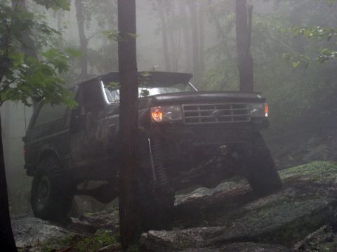 1988 Ford Bronco - A dark, mud-splattered Ford Bronco climbs a wet granite ledge in dense fog, headlights and marker lights on and the driver's-side front tir…