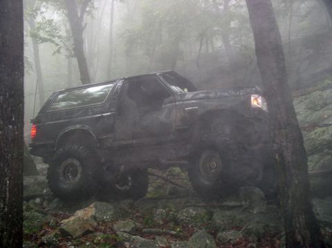1988 Ford Bronco - Side view of a mud-caked lifted Ford Bronco with its doors removed and hardtop still on, flexing up a steep rock ledge in fog-filled woods.…
