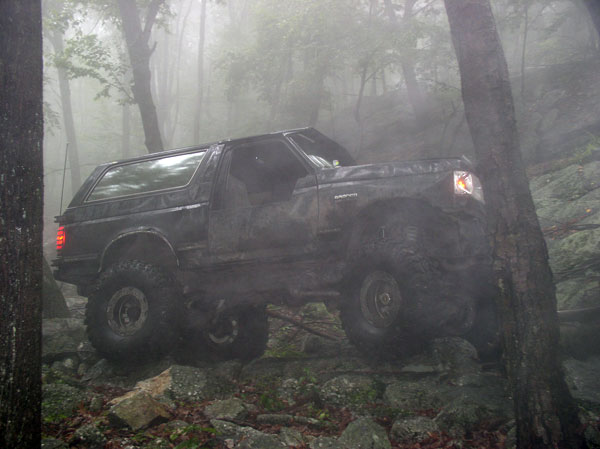 Muddy lifted Ford Bronco with doors removed climbing a rock ledge in foggy woods