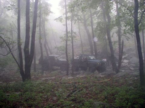 1988 Ford Bronco - Two off-road vehicles navigate a rocky, wooded area shrouded in fog.